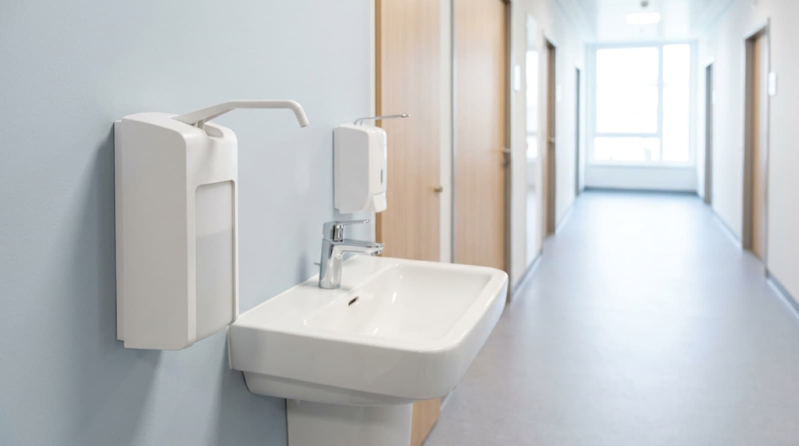 Weißer Wandspender für Händedesinfektion mit langem Bedienbügel neben einem weißen Handwaschbecken in einem hellen Praxisflur mit Holztüren