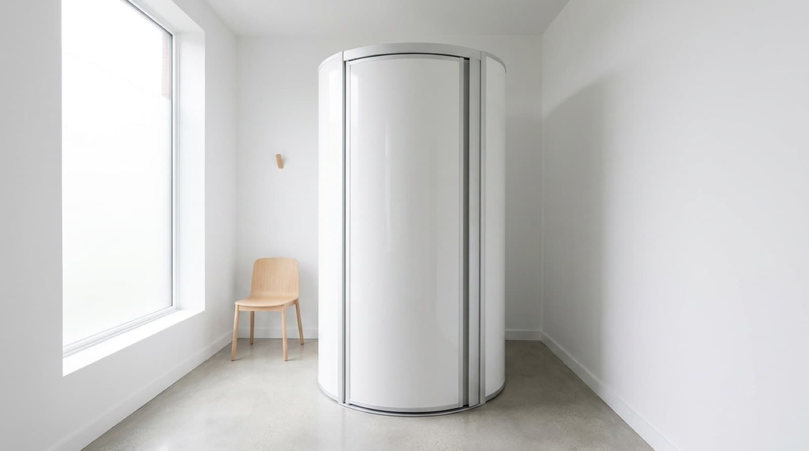 Heller, leerer Behandlungsraum mit einer geschlossenen weißen, runden Lichttherapie-Kabine, daneben ein Holzstuhl und ein großes Fenster