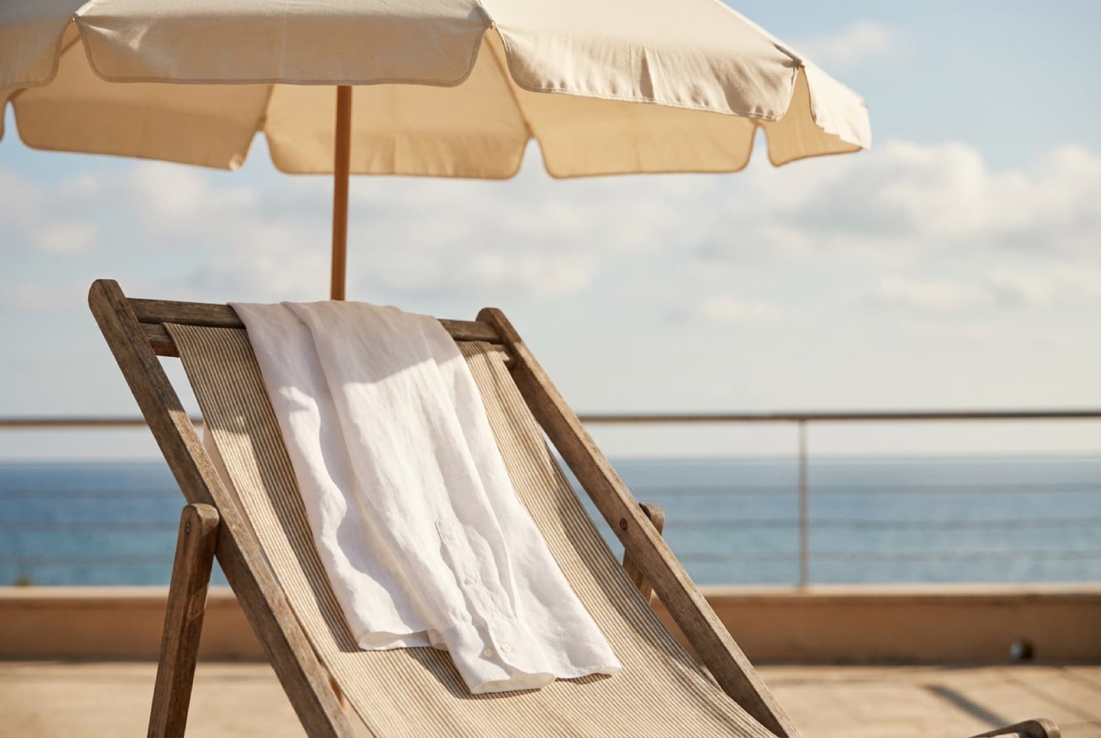 Heller Sonnenschirm über einem hölzernen Liegestuhl, auf dem ein langärmeliges weißes Leinenhemd liegt, im Hintergrund das Meer
