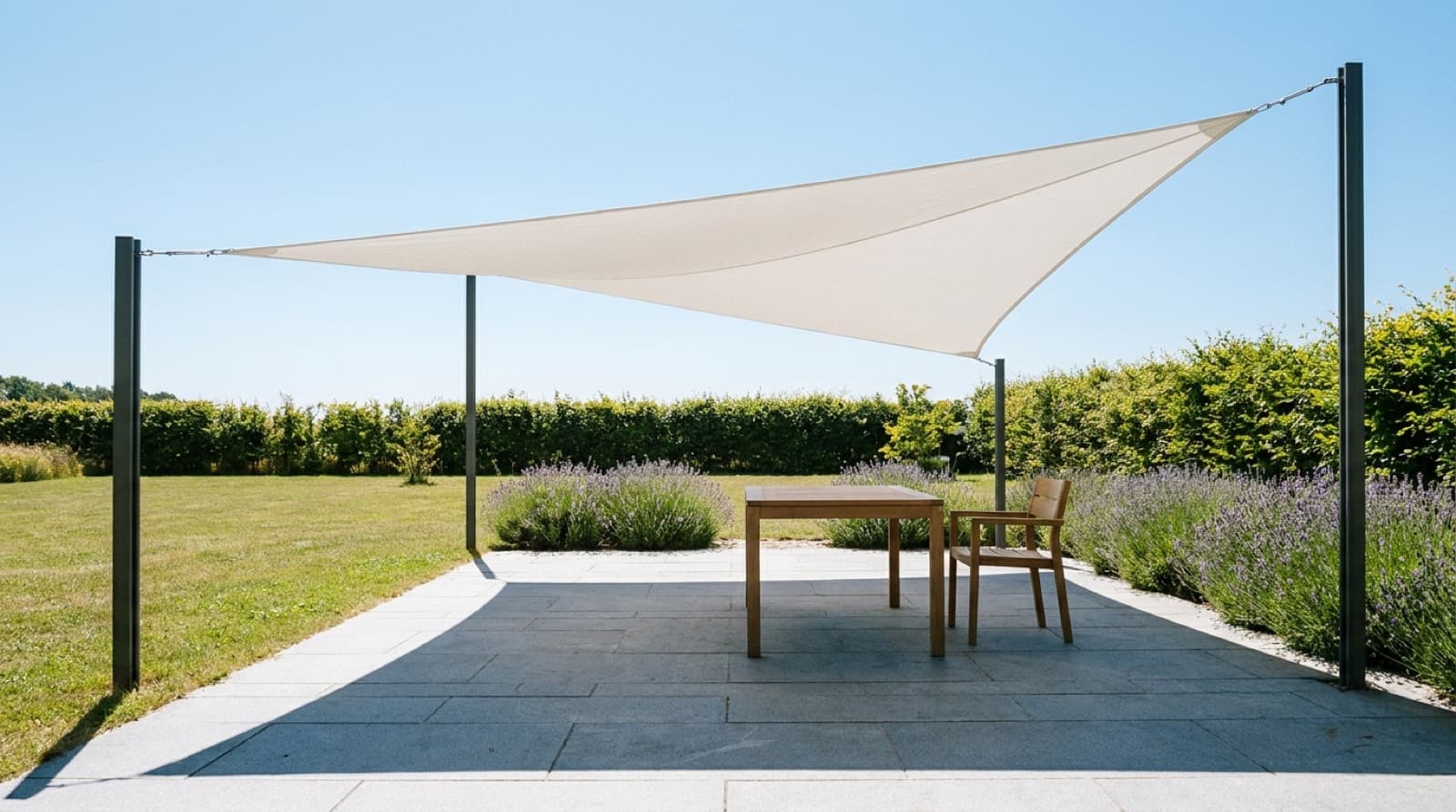 Großes weißes, dreieckiges Sonnensegel spannt sich über eine Gartenterrasse und wirft einen klaren Schatten auf einen leeren Holztisch mit Stuhl, dahinter Lavendel, Rasen und eine Hecke im Mittagslicht